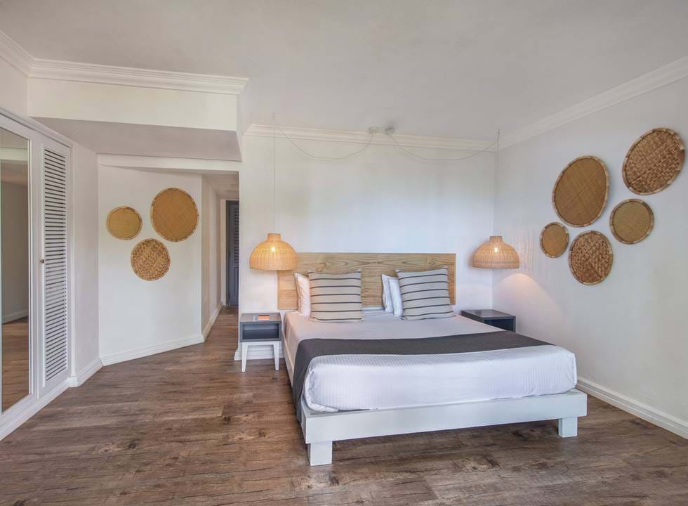 Moderní ložnice s dřevěnou podlahou, postelí s pruhovanými polštáři a dřevěným čelem, dvěma nočními stolky s proutěnými lampami a tkanými koši zavěšenými na stěnách.